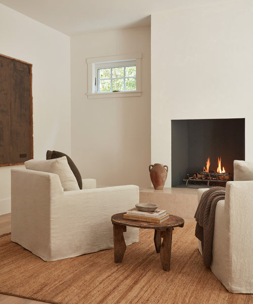 A cozy living room features two Harbor Chair Slipcovers in natural linen facing a modern fireplace, with a rustic wooden coffee table, books, a brown vase, a textured rug, and a small window on the cream wall.