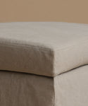 A close-up of a light beige upholstered ottoman corner, showing the fabric texture and stitched seam against a matching neutral background.