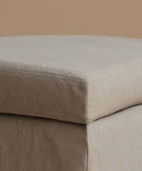 A close-up of a light beige upholstered ottoman corner, showing the fabric texture and stitched seam against a matching neutral background.