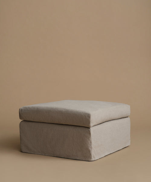 A square upholstered ottoman or stool cushion in a light taupe-beige fabric, shown against a solid warm beige studio background.