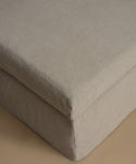 A close-up of a light beige upholstered ottoman or bench corner, showing the fabric texture and stitched seam against a matching neutral background.