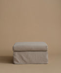 A square upholstered ottoman or stool cushion in a light taupe-beige fabric, shown against a solid warm beige studio background.