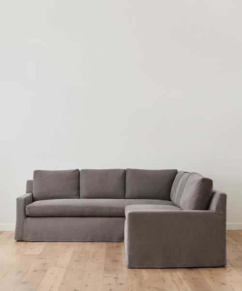 The Harbor Sectional, featuring Belgian Flax Linen upholstery, is arranged on a light wooden floor with a plain white wall backdrop in a minimalist room.