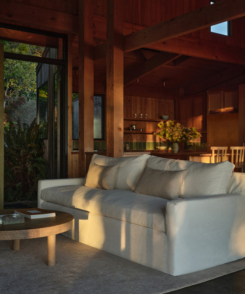 A cozy living room features the Harbor Sofa in ivory linen, a wooden coffee table, and wood-paneled walls with beams. Sunlight streams in, highlighting a dining area and greenery in the background. The Harbor Sofa is handmade in Los Angeles.