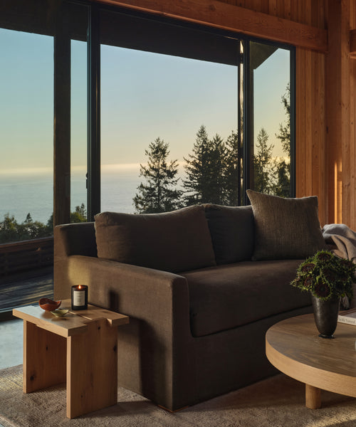 A cozy living room showcases a Harbor Sofa with linen upholstery, wooden tables, and large windows framing sunset views of trees and the sea. A candle and bowl sit on the side table, while a plant adorns the coffee table.