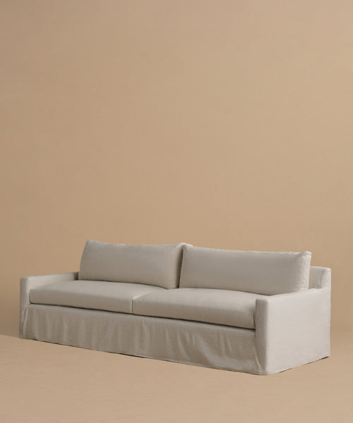 The Harbor Sofa, handmade in Los Angeles, features a slipcover, two seat cushions, and clean lines in natural linen. Set against a solid beige background, it embodies a modern, minimalist style.