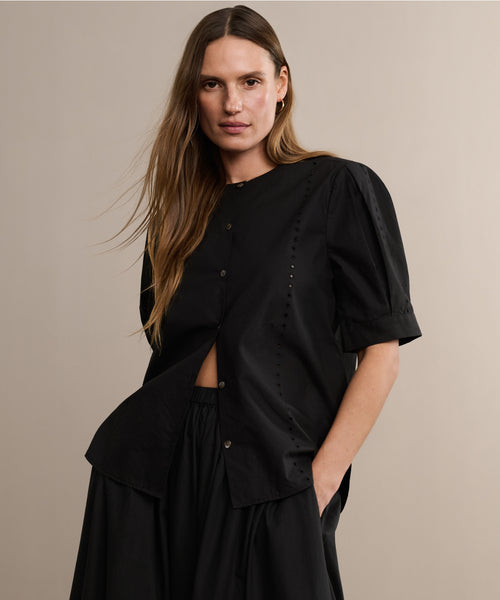 A woman with long brown hair wears the Harper Blouse in black Italian cotton poplin with short sleeves and a matching skirt, standing against a plain beige background.