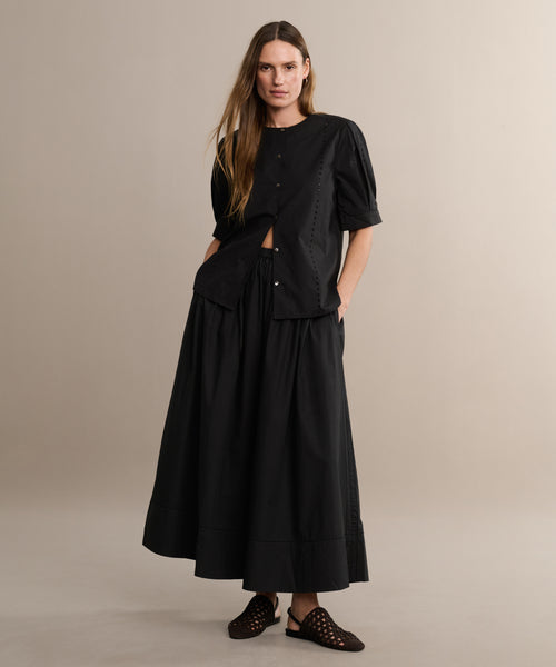 A woman with long hair stands in a studio, wearing a loose-fitting Harper Blouse and a matching flared skirt in Italian cotton poplin. Her hands are in her pockets and she wears black woven slip-on shoes against a plain background.