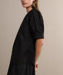 A woman models the Harper Blouse in black Italian cotton poplin with embroidered eyelets, paired with a matching black skirt. She stands against a beige background, her face partially out of frame.