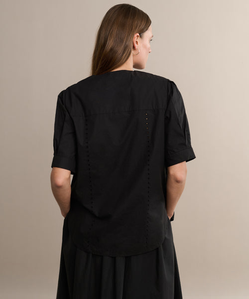 A woman with long brown hair wears a loose black Harper Blouse and a matching skirt, standing with her back to the camera against a plain beige background.