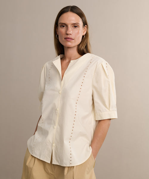 A woman with straight brown hair wears the Harper Blouse, featuring embroidered eyelets, short sleeves, and a buttoned front in Italian cotton poplin. She pairs it with beige pants and stands against a light tan background.