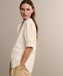 A woman with long brown hair wears the Harper Blouse and tan pants, standing sideways against a neutral beige background with a soft, serene expression.