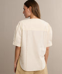 A woman with straight brown hair models the Harper Blouse, a cream Italian cotton poplin top with short sleeves and embroidered eyelets at the back seams. She stands with her back to the camera against a neutral background.