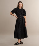 A woman with straight brown hair and a neutral expression wears the Harper Dress—an Italian cotton poplin, black button-down short-sleeve midi with a fitted waist—and black sandals, standing against a plain beige background.
