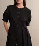 A woman wearing the Harper Dress—a black, short-sleeved Italian cotton poplin button-down with puffed shoulders—stands against a neutral background. Her face is partially out of frame.