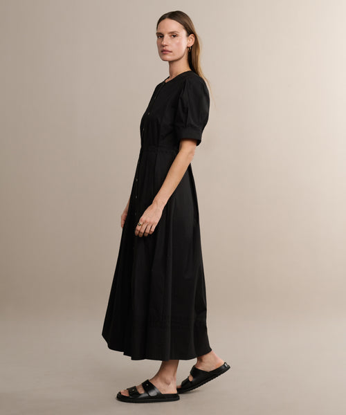 A person stands sideways in the Harper Dress with short puffed sleeves and black sandals, set against a plain beige backdrop.