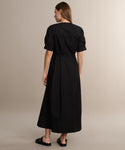 A woman with straight brown hair faces away, wearing the Harper Dress in black Italian cotton poplin and black slip-on sandals, set against a plain beige background.
