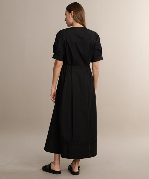 A woman with straight brown hair faces away, wearing the Harper Dress in black Italian cotton poplin and black slip-on sandals, set against a plain beige background.