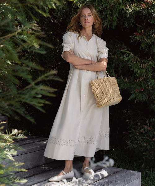 A woman stands outdoors amid lush greenery, wearing a cream Harper Dress and white shoes, holding a woven straw bag with her arms crossed.