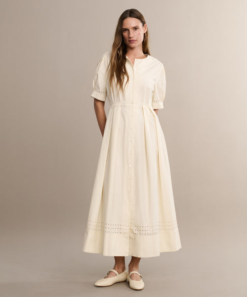 A woman stands in a neutral studio wearing the Harper Dress—a cream, button-down Italian cotton poplin midi with embroidered eyelets at the hem and short puff sleeves—paired with neutral flats.