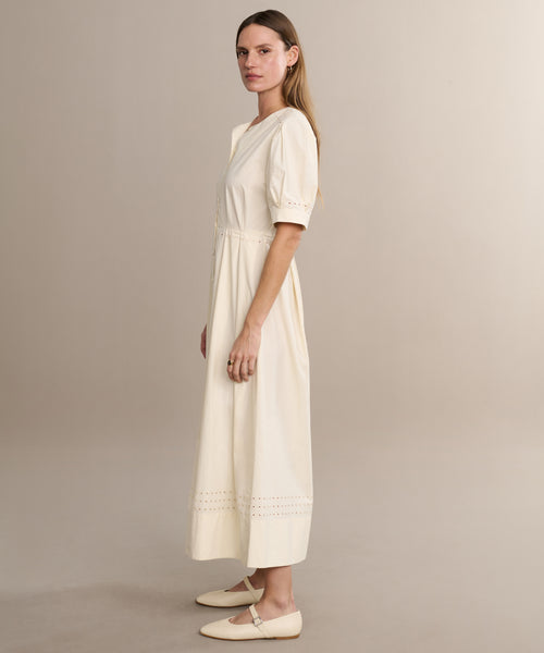 A woman stands sideways wearing the Harper Dress, featuring embroidered eyelets near the hem, paired with cream flats against a plain beige background.