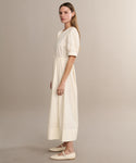 A woman in profile wears the Harper Dress—an ankle-length, cream Italian cotton poplin piece with subtle embroidered eyelets and matching cream flats—set against a plain beige backdrop.
