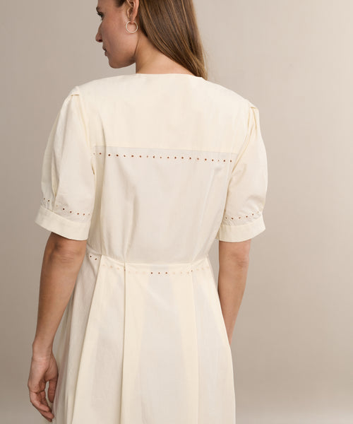 A woman with long brown hair wears the Harper Dress in cream Italian cotton poplin with embroidered eyelets on the back yoke and sleeves, standing with her back to the camera against a beige background.