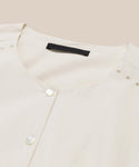 A close-up of the Harper Dress in cream Italian cotton poplin features a round neckline, two white buttons, delicate embroidered eyelets by the shoulders, and a black label inside the collar, set against a beige background.