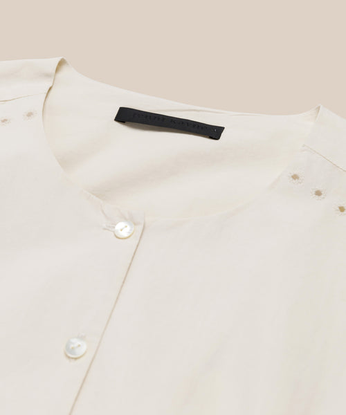 A close-up of the Harper Dress in cream Italian cotton poplin features a round neckline, two white buttons, delicate embroidered eyelets by the shoulders, and a black label inside the collar, set against a beige background.