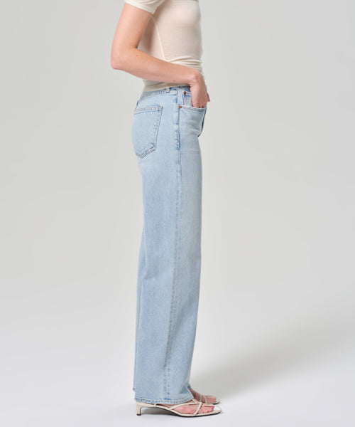 A person in profile wears a fitted short-sleeve beige top, white heeled sandals, and the Harper Jean: light blue organic cotton jeans with a wide-leg silhouette, set against a plain light background.