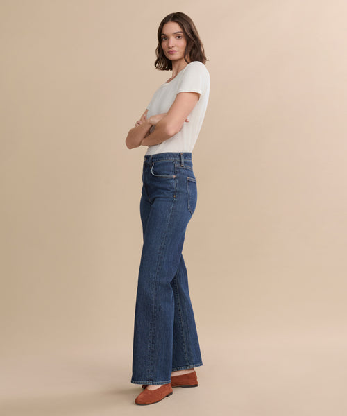 A person stands sideways with arms crossed, wearing a white short-sleeve shirt, the Harper Jean in an organic cotton wide-leg silhouette, and brown shoes against a plain beige background.