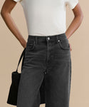 Wearing a plain white T-shirt tucked into Harper Jean’s high-waisted, wide-leg denim, a person stands with one hand on their hip holding a black bag, the modern silhouette set against a solid beige background.