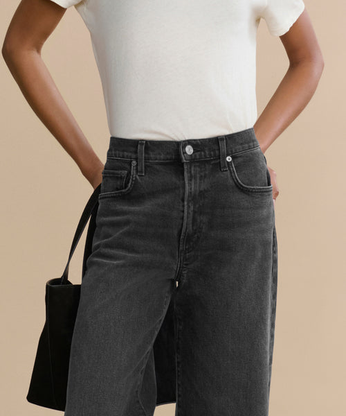 Wearing a plain white T-shirt tucked into Harper Jean’s high-waisted, wide-leg denim, a person stands with one hand on their hip holding a black bag, the modern silhouette set against a solid beige background.