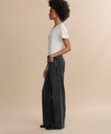 A person with curly hair stands in profile against a beige background, wearing a white T-shirt, the Harper Jean—relaxed mid-rise organic cotton jeans in a wide-leg silhouette—and black shoes, with one hand in their pocket.