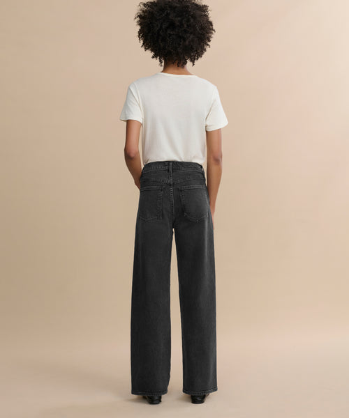 A person with curly hair faces away, wearing a white short-sleeve t-shirt and the Harper Jean—relaxed mid-rise, wide-leg jeans—against a plain beige background.