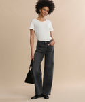 A woman stands against a beige background, wearing a white short-sleeve shirt and the Harper Jean—relaxed mid-rise, wide-leg jeans. She finishes the look with black shoes, a black bag, and a slight smile as her curly hair frames her face.
