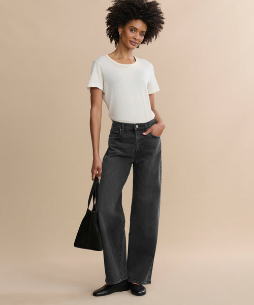 A woman stands against a beige background, wearing a white short-sleeve shirt and the Harper Jean—relaxed mid-rise, wide-leg jeans. She finishes the look with black shoes, a black bag, and a slight smile as her curly hair frames her face.