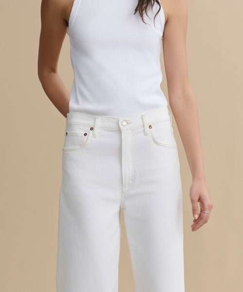 A person wearing a white sleeveless top and high-waisted Harper Jean stands against a beige background, with one hand behind their back and the other relaxed by their side.