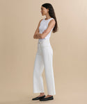A woman with long dark hair stands in profile against a beige background, wearing a white sleeveless top and Harper Jean—high-waisted organic cotton jeans with a wide-leg silhouette. She pairs them with classic black flats and has her arms crossed as she looks ahead.