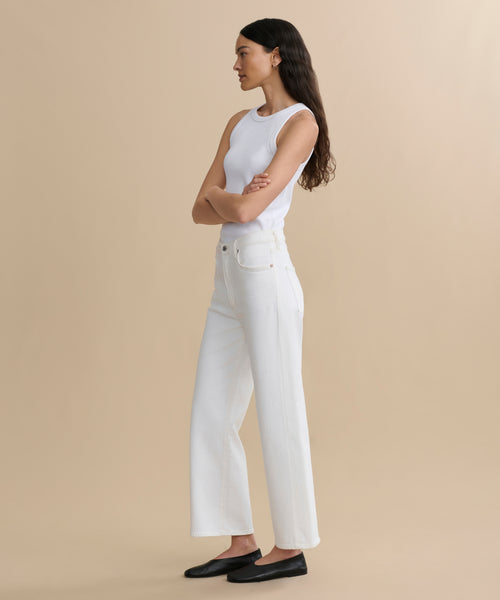 A woman with long dark hair stands in profile against a beige background, wearing a white sleeveless top and Harper Jean—high-waisted organic cotton jeans with a wide-leg silhouette. She pairs them with classic black flats and has her arms crossed as she looks ahead.