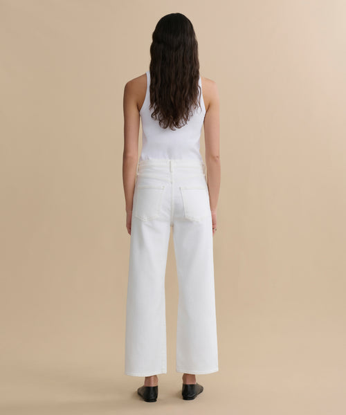 A woman with long wavy brown hair, in a white sleeveless top, Harper Jean wide-leg white wash jeans, and black flats, stands facing away against a plain beige background.