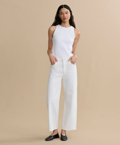 A woman with long dark hair stands against a beige background, wearing the high-waisted Harper Jean in organic cotton with a wide-leg silhouette, a white sleeveless top, and black flats. Her hands are in her pockets.