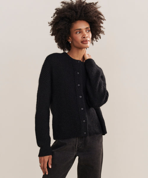 A person with short, curly hair wears the black Hayes Crewneck Cardigan and dark jeans, standing against a plain light background with one hand near their neck and a relaxed expression.