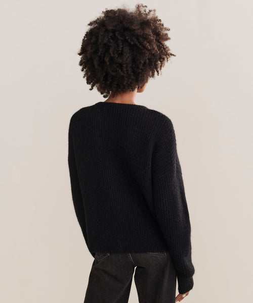 A person with short, curly hair stands with their back to the camera, wearing the Hayes Crewneck Cardigan in black and dark pants against a plain, light-colored background.