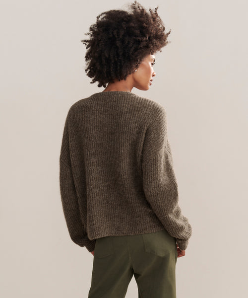 A person with curly hair, seen from behind, is wearing the Hayes Crewneck Cardigan and olive green pants, standing against a plain beige background.