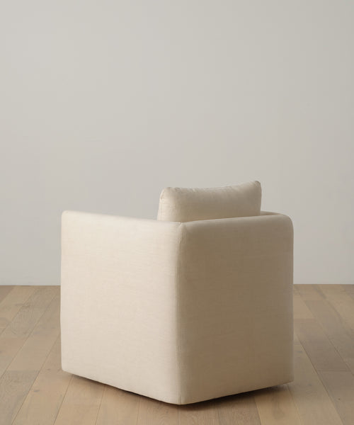 The Dume Chair, a modern minimalist accent chair in Beige Hemp Plush with clean square lines, sits on a light wood floor against an off-white wall. Handmade in Los Angeles, it’s shown from a rear side angle.