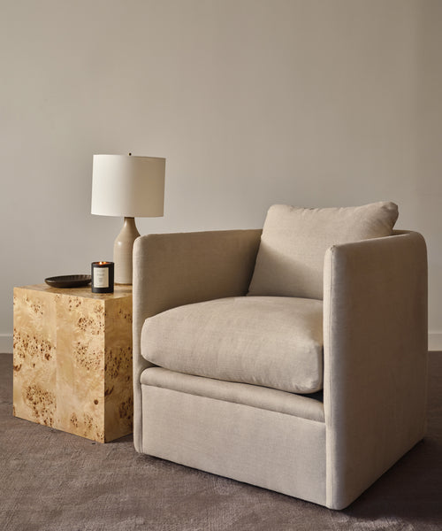 The Dume Chair in beige hemp stands beside a square wooden side table holding a lamp, candle, and small dish on a carpeted floor, offering sustainable style against a plain light-colored wall.