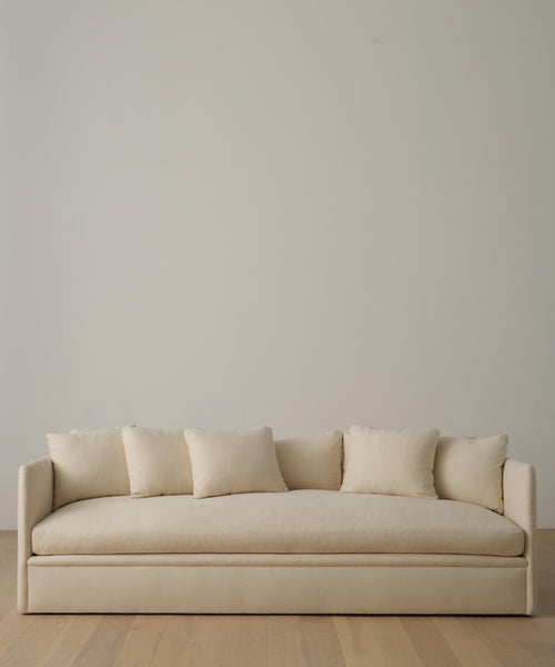 The Dume Sofa in beige hemp, with five matching cushions, stands against a light wall on a pale wooden floor, featuring eco-friendly, sustainable hemp upholstery for a minimalist touch.