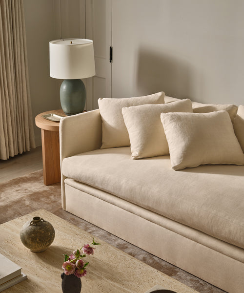 The beige Dume Sofa with matching cushions sits by a wooden side table topped with a green lamp in a neutral living room. On the stone coffee table in front, a ceramic vase holds pink flowers.