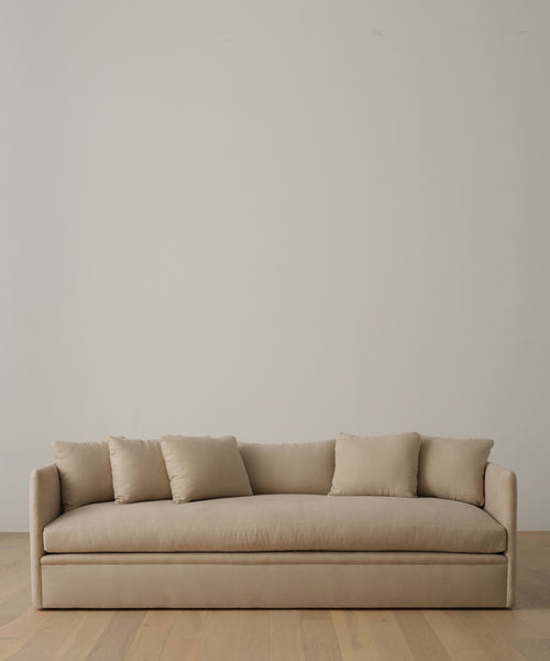 The Dume Sofa, a minimalist beige hemp piece handmade in Los Angeles with sustainable upholstery, includes four matching cushions and sits against an off-white wall on a light wooden floor.